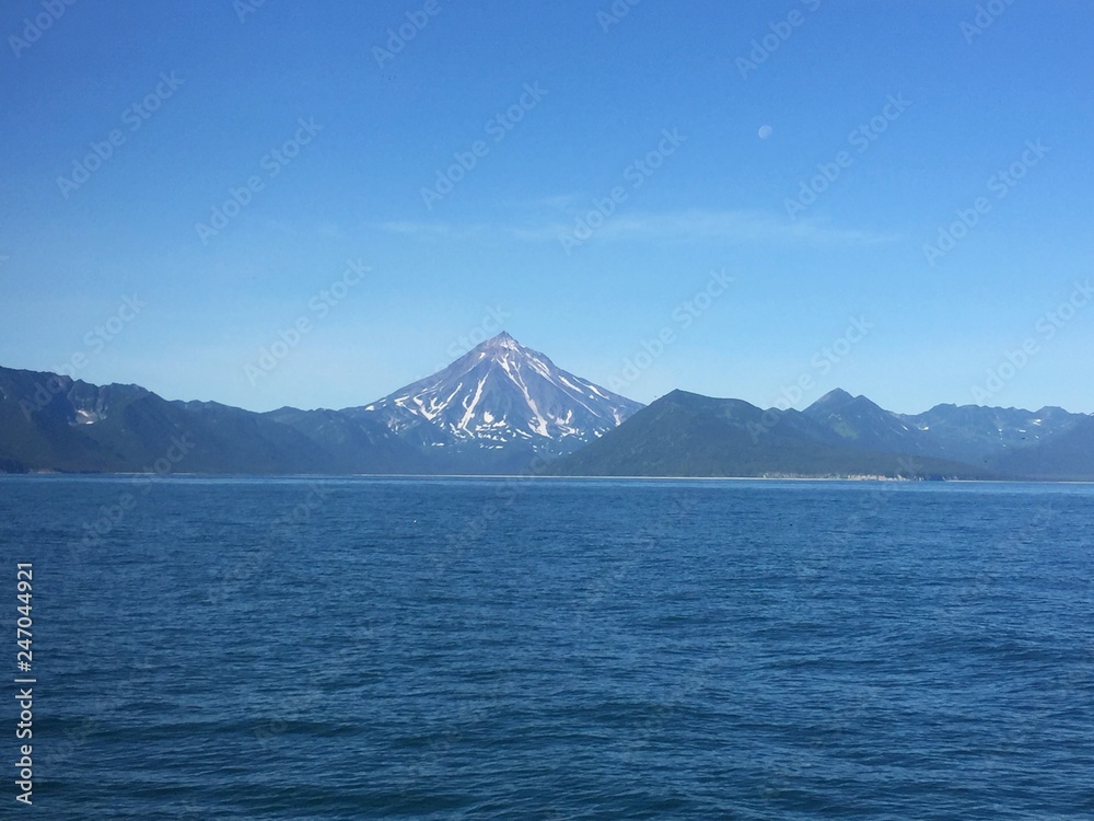View of Vilyuchinsky volcano (also called Vilyuchik) from water. It's a stratovolcano in the southern part of Kamchatka Peninsula, Russia. Moon is visible in the sky.