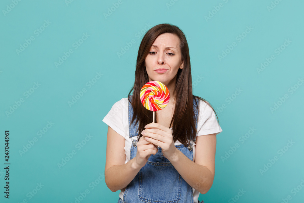 Portrait of puzzled displeased young woman in denim clothes hold, looking on colorful round lollipop isolated on blue turquoise wall background in studio. People lifestyle concept. Mock up copy space.