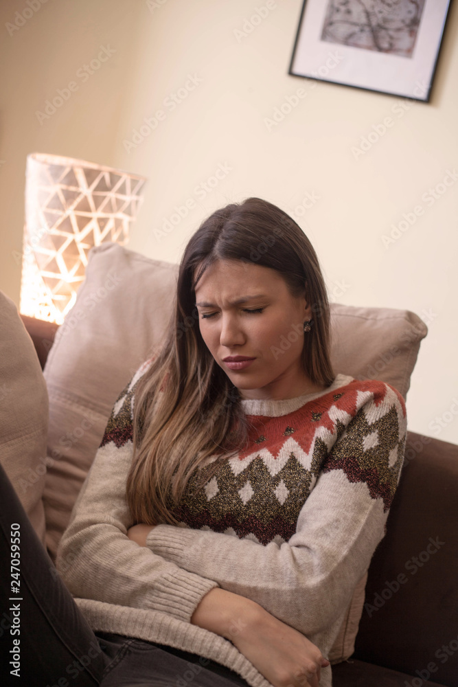 Unhealthy young woman with stomachache leaning on the couch at home.