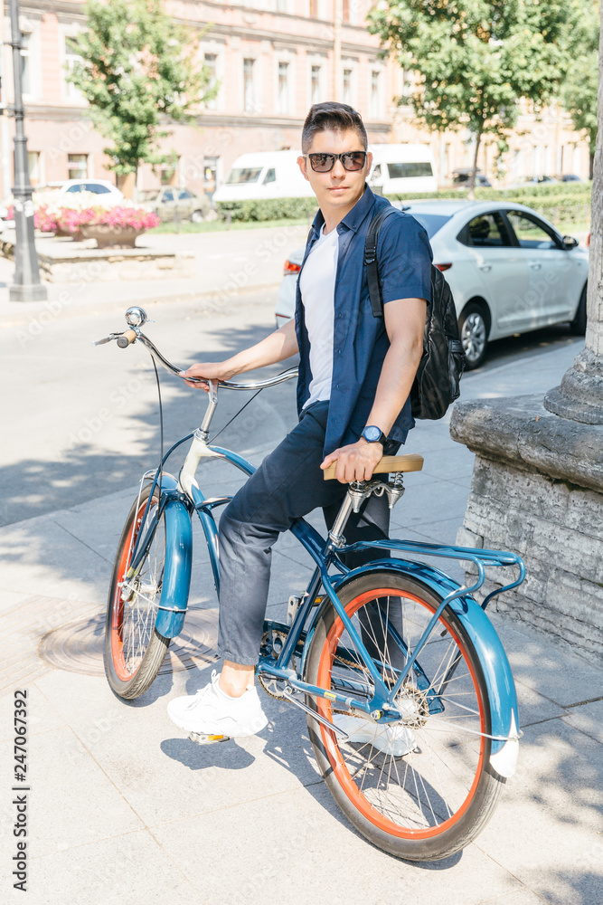 Young attractive man in sunglasses on the vintage blue bike wait green light/ outdoor portrait of man dressed in fashionable clothes/ handsome stylish guy bicycling in the city/ sport concept.