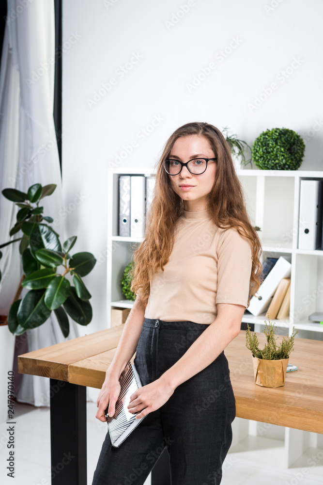 Portrait of a young Caucasian woman with long hair and glasses for friction stands in an office inside an office room near a desk. University student girl. Topic man learning Portrait of a young Caucasian woman with long hair and glasses for friction stands in an office inside an office room near a desk. University student girl. Topic man learning