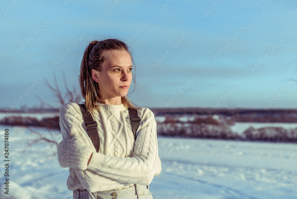 Portrait of a young girl in a white sweater and mittens. In winter, the country looks into the distance in casual clothes. Happy woman resting on the nature during winter day. Free space for text. Portrait of a young girl in a white sweater and mittens. In winter, the country looks into the distance in casual clothes. Happy woman resting on the nature during winter day. Free space for text.