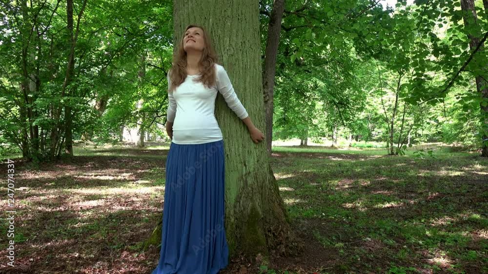 Relaxed pregnant woman standing lean on big tree trunk in park Relaxed pregnant woman standing lean on big tree trunk in park