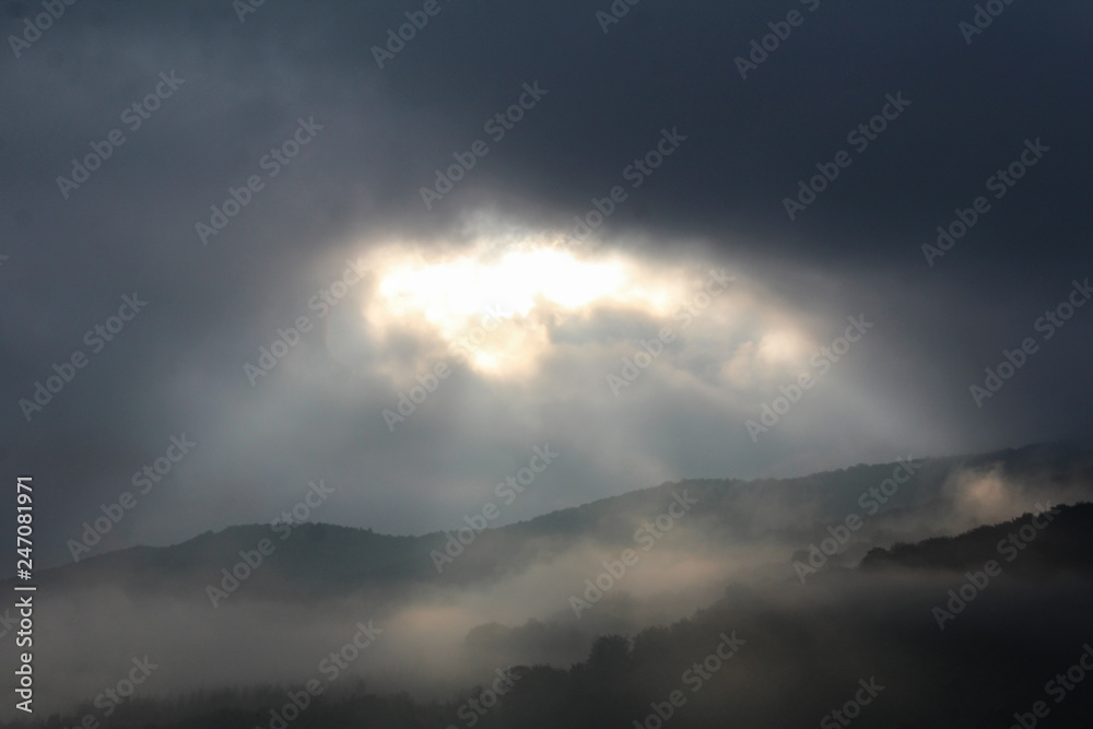 Sunbeams push through storm clouds, lighting up the mountain forest of Carpathians in fog..