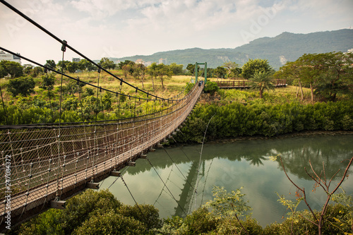 Narrow walking bridge suspended by cables over river between lush green landscape