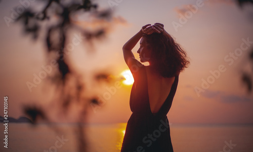 Young beautiful sensual woman in a stylish dress with an open back is standing on the seashore at sunset in the rays of the sun, beauty and fashion, leisure and travel concept