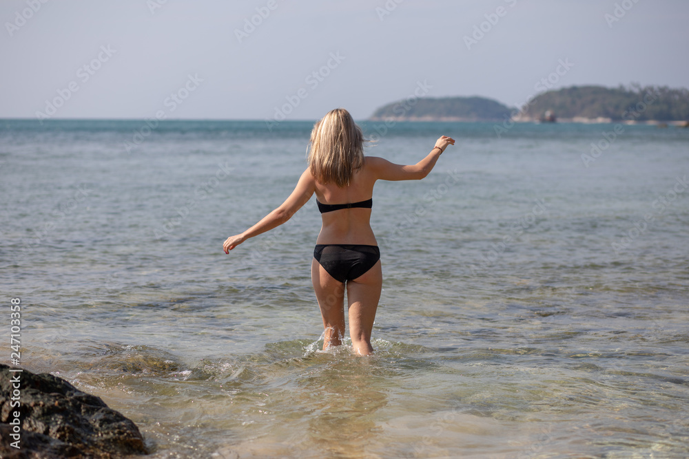 Young blonde woman in a black bikini looking at sea water on the beach of tropical resort. Trips to warm destinations. Phuket. Thailand. Rear view. Young blonde woman in a black bikini looking at sea water on the beach of tropical resort. Trips to warm destinations. Phuket. Thailand. Rear view.