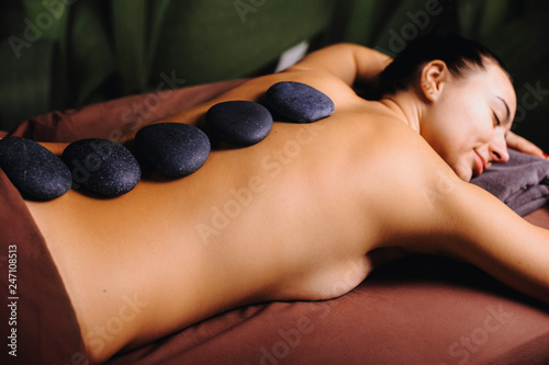 Close up of a young woman doing stone therapy with hot stones lying on bed with closed eyes. Body of female with spa black stones on it.