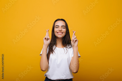 Portrait of adorable hopeful woman with crossed fingers , prays and whishes for good luck with closed eyes isolated over yellow background. photo