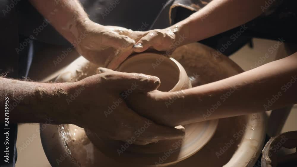 Two intertwined hands form a pot of clay on a potter's wheel. A male hand helps a woman's hands shape a clay shape.