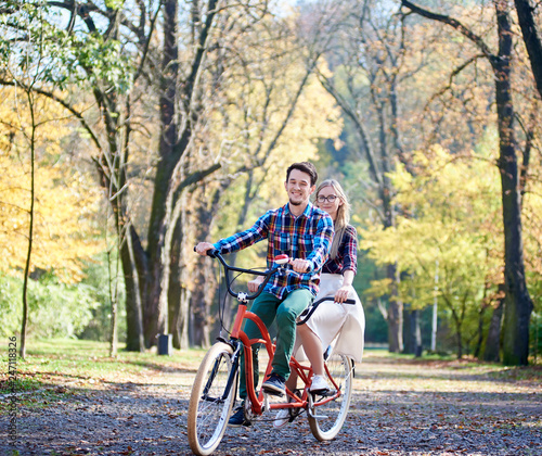 Young active couple, handsome bearded man and attractive blond long-haired woman cycling together tandem double bike by sunny alley with golden leaves on tall trees background