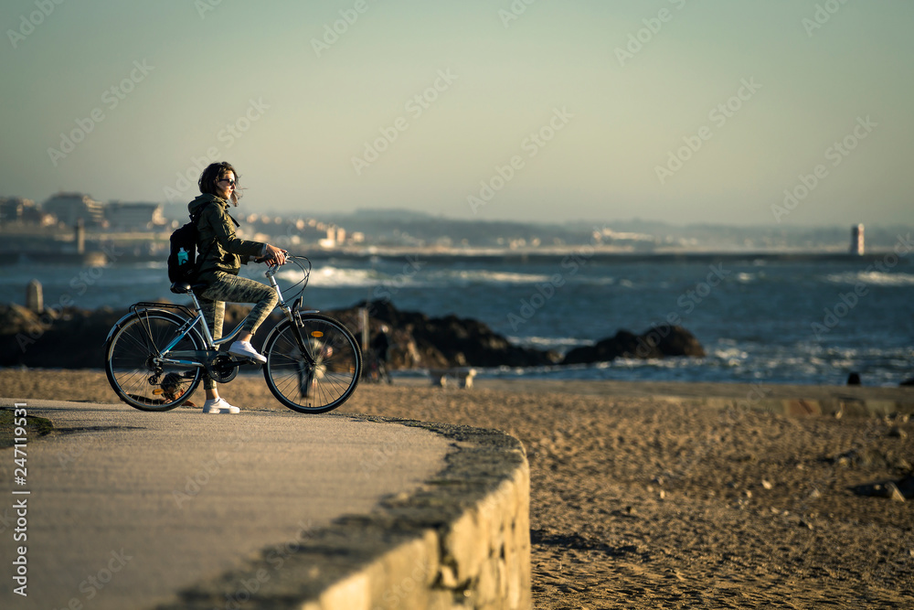 Young and beautiful girl is riding a bike by pier next to the Atlantic ocean during sunset time. Beautiful light. Smiley face. Porto, Portugal.