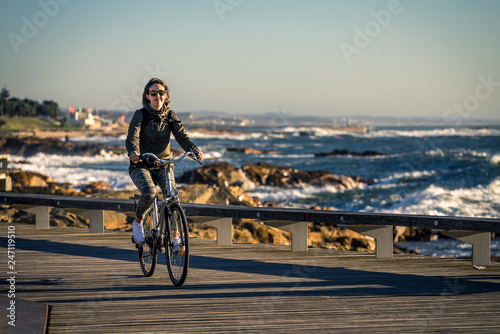 Young and beautiful girl is riding a bike by pier next to the Atlantic ocean during sunset time. Beautiful light. Smiley face. Porto, Portugal.