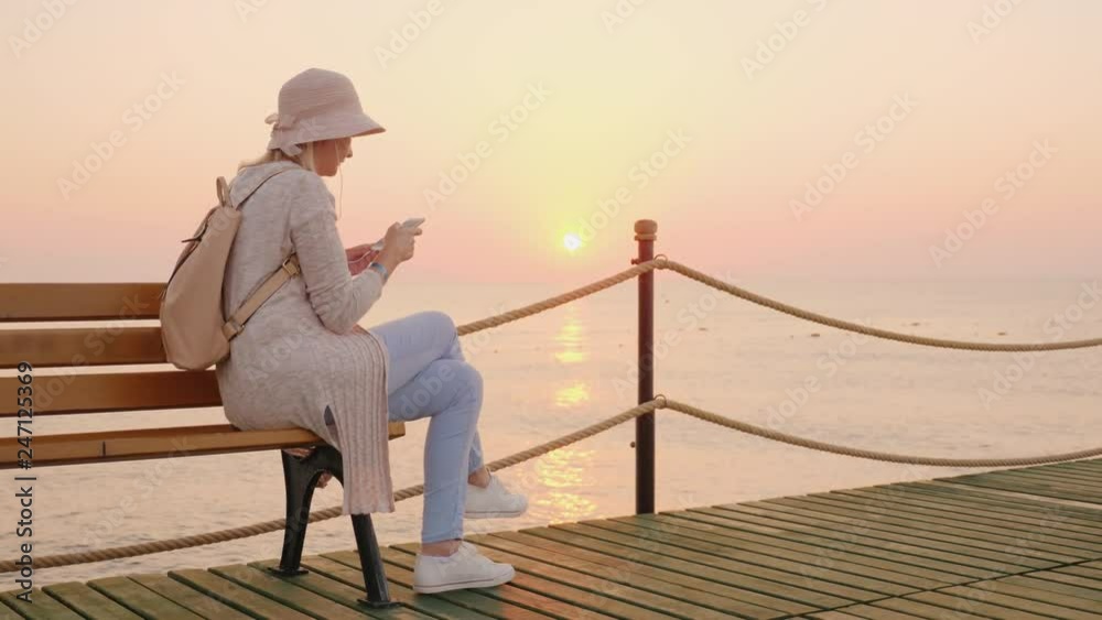A stylish girl in a pink hat sits on a bench on a sea pier and reads a text message on the phone, the rising sun is reflected in the blue water, coloring the sky in pink A stylish girl in a pink hat sits on a bench on a sea pier and reads a text message on the phone, the rising sun is reflected in the blue water, coloring the sky in pink