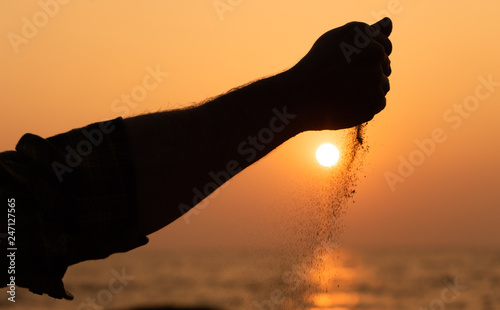  a man sits at sunset on the shore of the ocean and pours sand from his hands against the background of the sun and sky