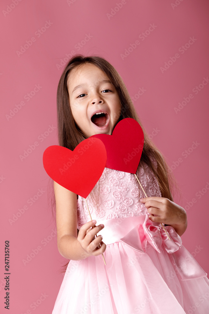laughing cute little girl in a pink dress holding two paper heart on pink background. St. Valentine's Day laughing cute little girl in a pink dress holding two paper heart on pink background. St. Valentine's Day