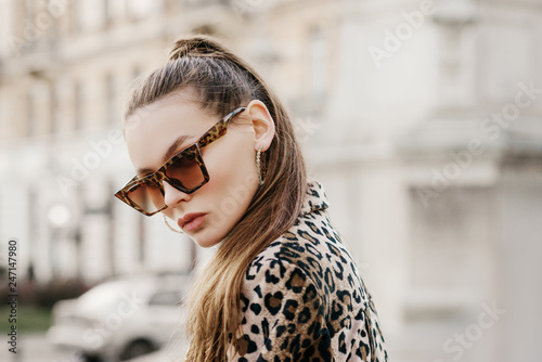  Outdoor close up fashion portrait of young beautiful fashionable woman wearing stylish animal, leopard print sunglasses, hoop earrings, blazer, walking in street of european city. Copy, empty space