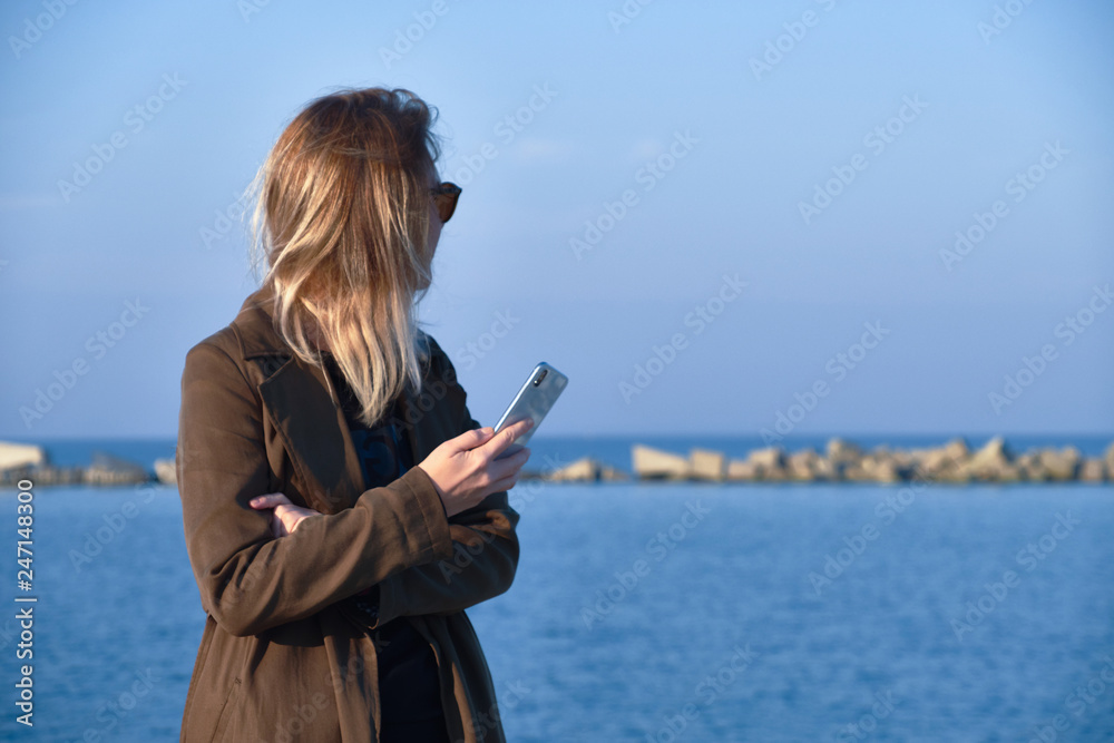 Naklejka premium Charming blonde girl with a phone, standing on the pier