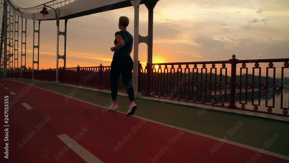 woman athlete running in city at sunrise. girl with short haircut training cardio runs on the bridge slow summer season slow motion woman athlete running in city at sunrise. girl with short haircut training cardio runs on the bridge slow summer season slow motion