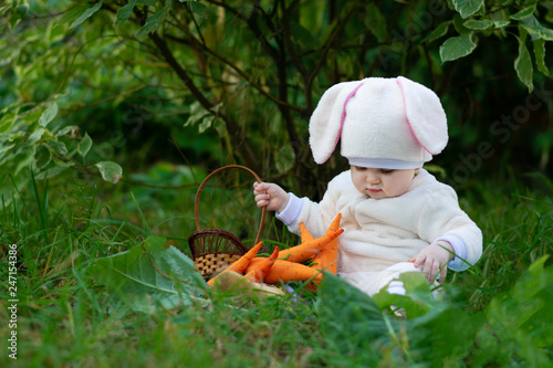 Baby girl in bunny costume seating on grass in forest and playing with toy carrots. © Vector DSGNR
