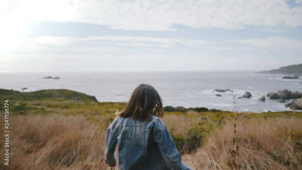 Camera follows young woman walking down on Big Sur ocean coast covered with yellow flowers, smiling cheerfully at camera Camera follows young woman walking down on Big Sur ocean coast covered with yellow flowers, smiling cheerfully at camera