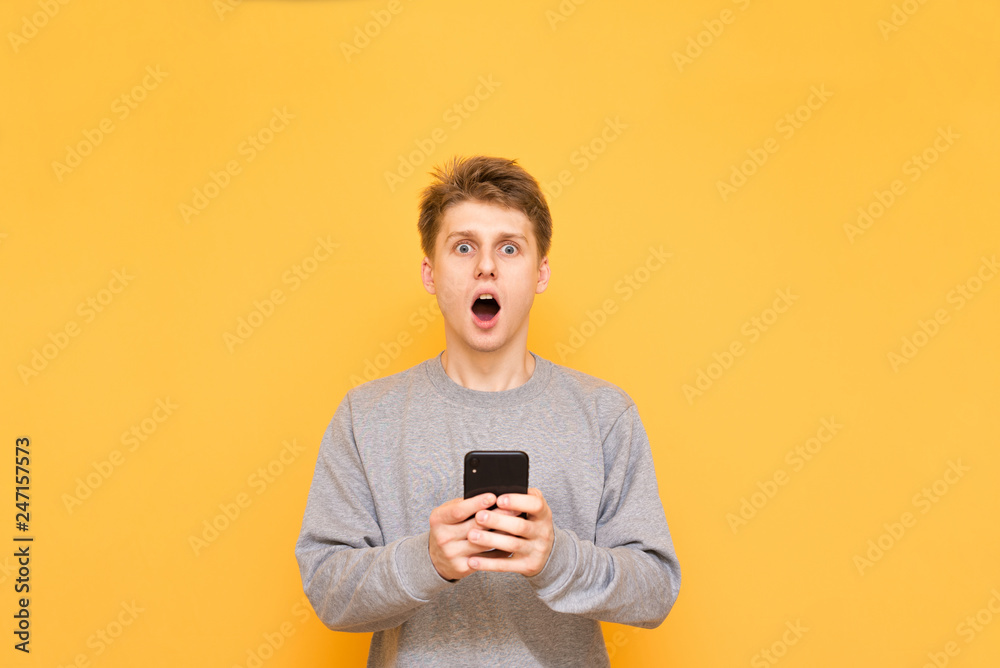 Expressive guy on a yellow background holds the smartphone in his hands, looking at the camera with astonished gaze. Amazed young man uses a smartphone isolated on a yellow background. Copyspace