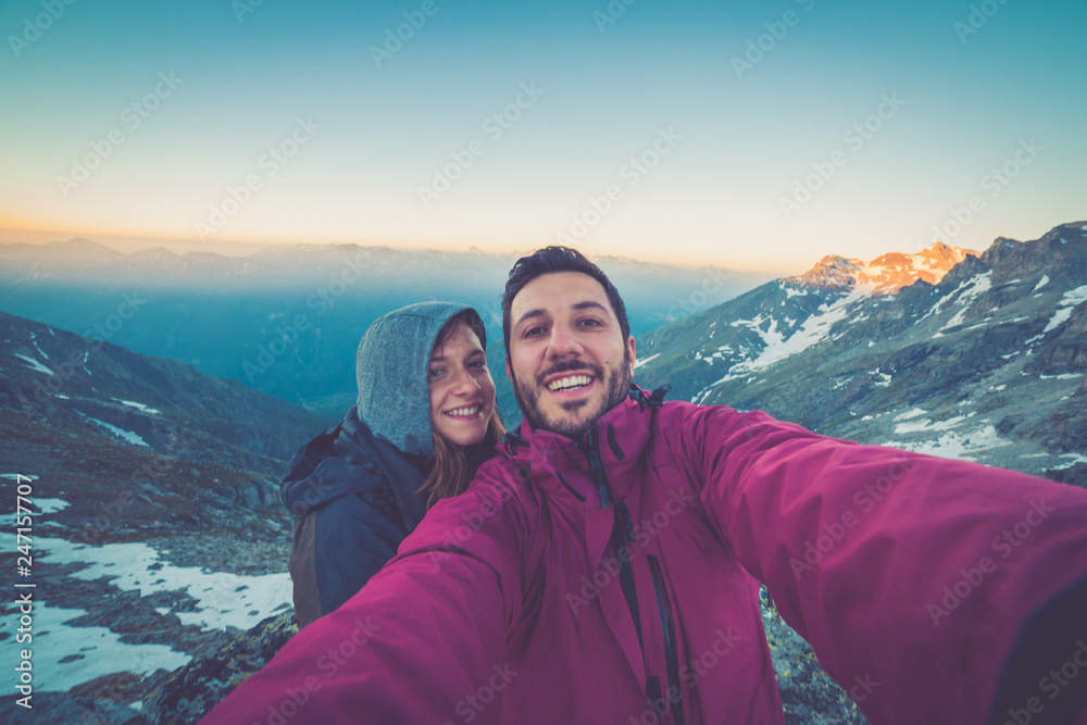 Couple of hikers taking selfie from top of the mountain with peaks and valley view on the background  after long trekking