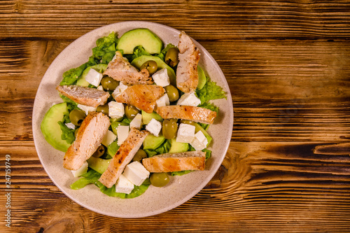 Fresh salad with chicken meat, feta cheese, avocado, green olives and lettuce leaves in ceramic plate on wooden table. Top view
