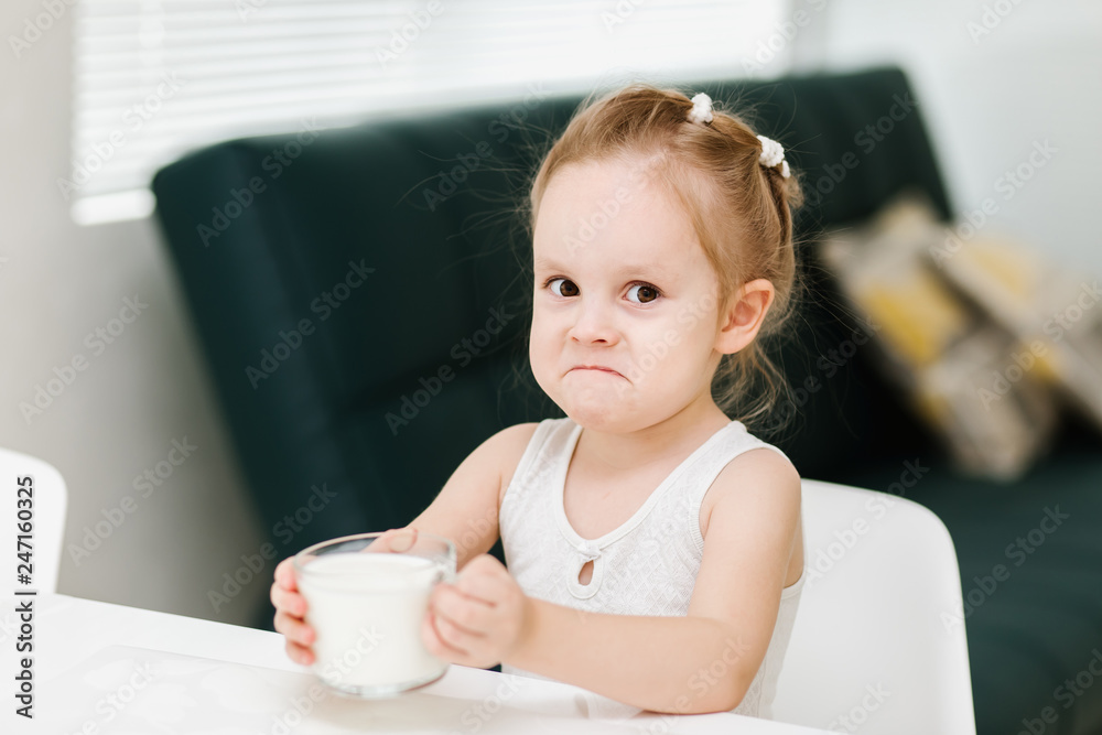 Little smart caucasian girl drinks milk from a glass cup in the kitchen and has fun, healthy eating concept for children. Little smart caucasian girl drinks milk from a glass cup in the kitchen and has fun, healthy eating concept for children.
