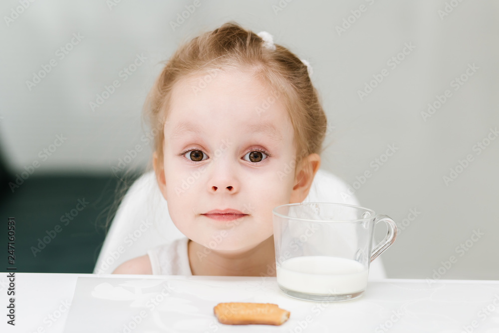 Little smart caucasian girl drinks milk from a glass cup in the kitchen and has fun, healthy eating concept for children. Little smart caucasian girl drinks milk from a glass cup in the kitchen and has fun, healthy eating concept for children.