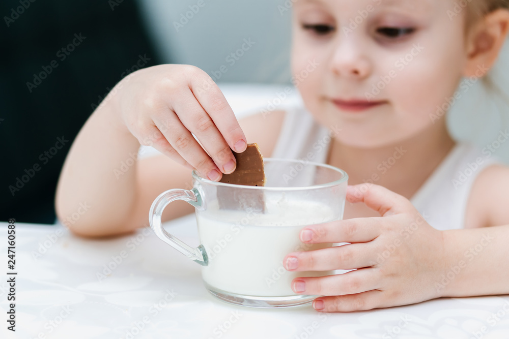 A little clever Caucasian girl drinks milk from a glass cup in the kitchen, wets chocolate cookies in a glass and has fun, healthy food for children. A little clever Caucasian girl drinks milk from a glass cup in the kitchen, wets chocolate cookies in a glass and has fun, healthy food for children.