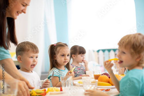 Kids and carer together eat fruits and vegetables in kindergarten or daycare
