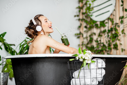 Young relaxed woman listening to the music and drinking smoothie while lying in the retro bathtub at the beautiful bathroom with green plants