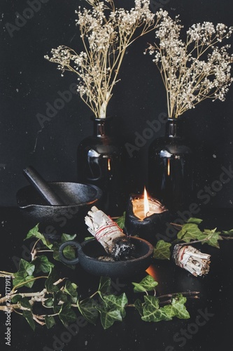 Vertical photo of sage sticks on wiccan witch altar. Two smudge sticks in cast iron holder. Ivy branches, dried flowers, black burning candle, mortar and pestle in background. White speckle overlay