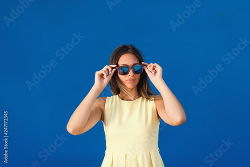 Portrait of beautiful Caucasian girl in light yellow dress putting on sunglasses while standing in front of blue background. © dusanpetkovic1