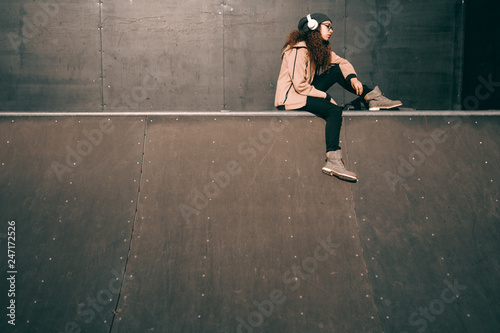 Mixed race hipster teenage girl with serious facial expression enjoying music and sitting in skate park.