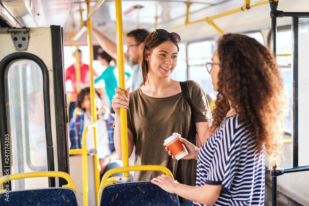 Two girls in a bus standing, chatting and smiling.