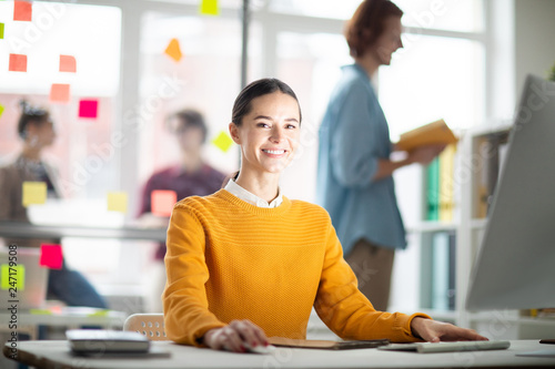 Pretty cheerful businesswoman in casualwear sitting by desk in front of computer monitor and carrying out her work