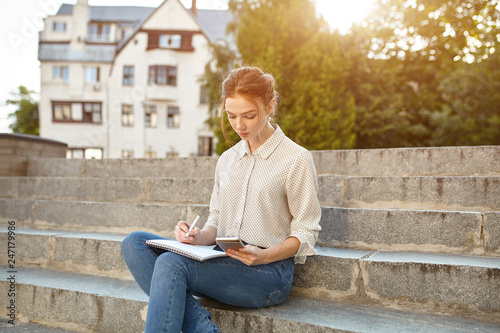 young beautiful student writes an essay in her notebook sitting on the steps stairs outdoor Red-haired girl with freckles