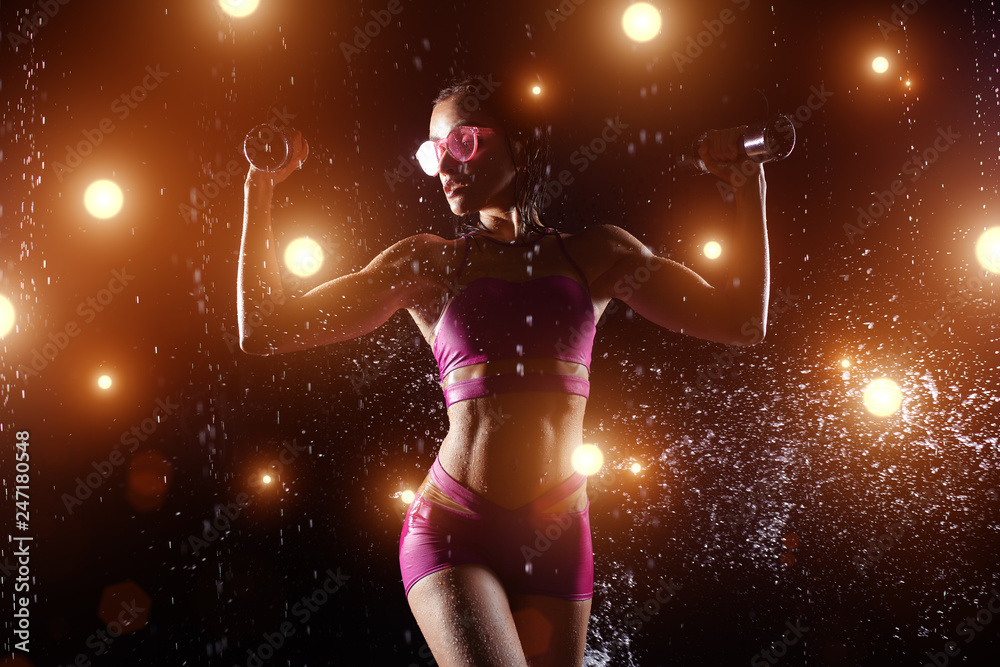 Beautiful sportswoman posing with dumbbells on a black background in spray of water