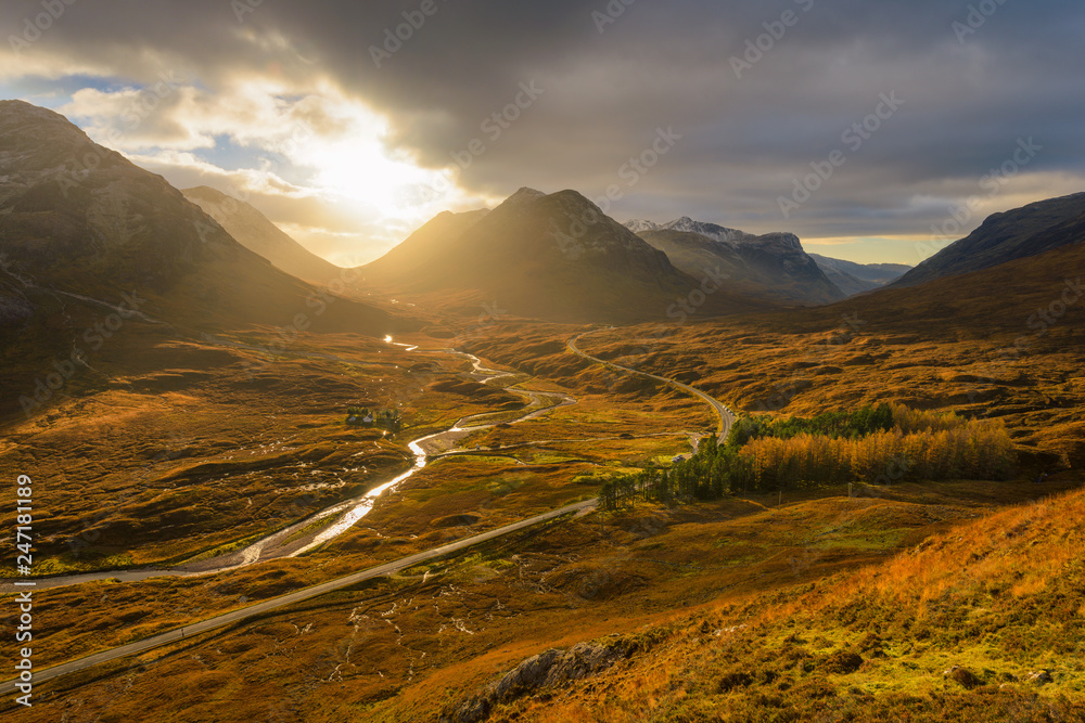 Winding Road And River Leading Through The Glencoe Valley With Golden Evening Light.
