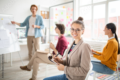 Young cheerful student with notepad making notes at seminar or training while teacher presenting important data photo