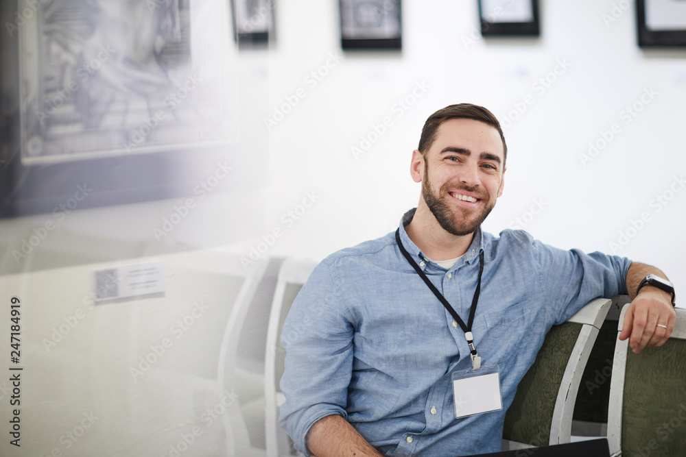 Portrait of smiling bearded man wearing blank name tag sitting on chair in art gallery or museum, copy space Portrait of smiling bearded man wearing blank name tag sitting on chair in art gallery or museum, copy space