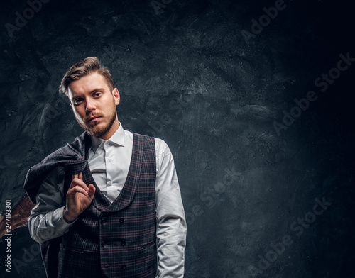 A confident elegantly dressed man holds jacket on the shoulder while posing against a dark textured wall