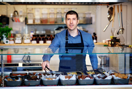 Young business owner standing behind the counter of his restaurant. Kitchen behind