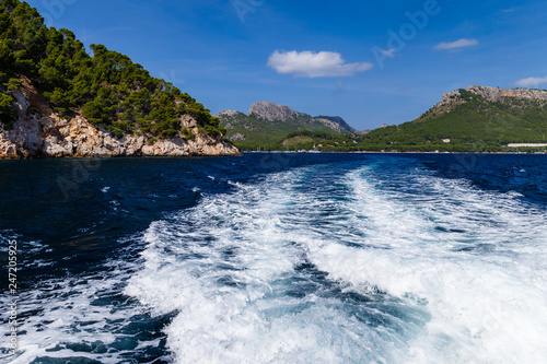 sunny day, sea view to the coast; view of the white waves left by the motorboat; on the shore there are large mountains partially overgrown with trees