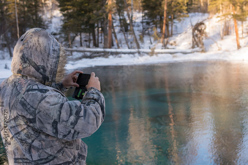 a man in camouflage winter clothes, taking pictures on a smartphone lake with a colorful turquoise bottom in the forest