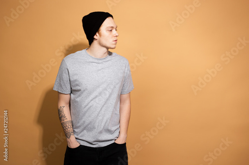 Stylish guy dressed in a gray shirt, black jeans and black hat is posing on the beige background in the studio