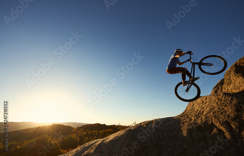 Cyclist standing on back wheel on trial bike. Professional sportsman rider making acrobatic stunt on the edge of big boulder on the top of mountain at sunset. Concept of extreme sport active lifestyle