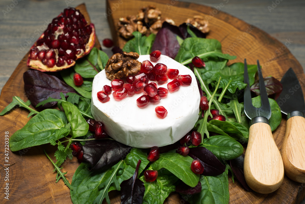Soft Camembert cheese, pomegranate grains and mix salad with young spinach, arugula, beet leaves on a brown wooden board. Tasty, gourmet food